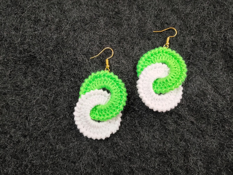 May include: A pair of crocheted earrings with a white and green swirl design. The earrings are made with a soft yarn and have gold hooks.