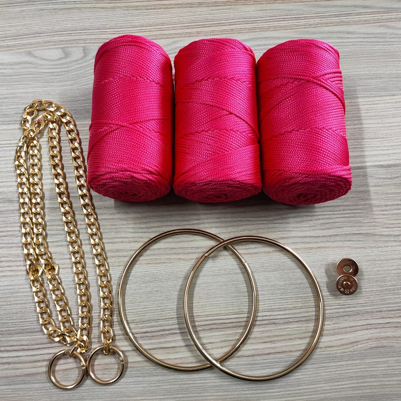 May include: Three cylindrical spools of vibrant hot pink cord, a long gold-tone metal chain with two attached rings, two large gold-tone circular metal hoops, and two small gold-tone circular metal fasteners with a central screw detail.