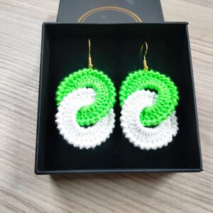 May include: A pair of green and white crocheted earrings in a black box. The earrings are in the shape of a circle with a smaller circle inside. The earrings are hanging from gold hooks.