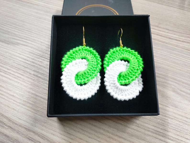 May include: A pair of green and white crocheted earrings in a black box. The earrings are in the shape of a circle with a smaller circle inside. The earrings are hanging from gold hooks.
