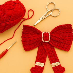 May include: A handmade red crochet bow with a white buckle and accents, alongside a ball of red yarn, a crochet hook, and small scissors. The bow is a festive craft item, perfect for embellishing gifts or decorations.