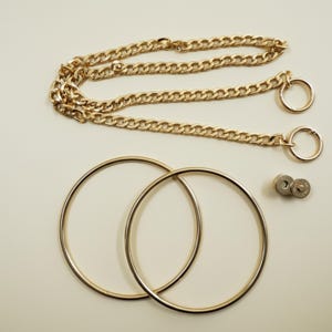 May include: A gold-toned metal bag accessory set featuring a long curb chain with two circular rings at the ends, two large smooth circular hoops, and a small circular magnetic snap closure button, all presented in a polished metallic finish.