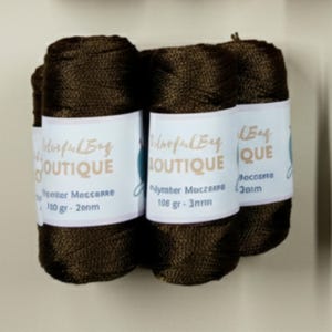 May include: Three spools of dark brown macrame cord wrapped in a woven texture. Each spool features a white paper label with the text ColorfulBag BOUTIQUE, Polyester Macrame, 108 gr, and 3mm printed in black and tan ink on the front surface.