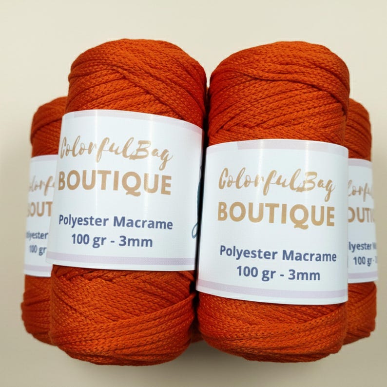 May include: Four cylindrical spools of vibrant orange polyester macrame cord. Each spool features a white paper label with the text Colorful Bag BOUTIQUE, Polyester Macrame, 100 gr - 3mm printed in dark brown and black ink on the front.
