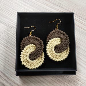 May include: A pair of crocheted earrings in a brown and beige color scheme. The earrings are shaped like a yin yang symbol and are hanging from gold hooks.