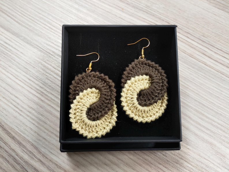 May include: A pair of crocheted earrings in a brown and beige color scheme. The earrings are shaped like a yin yang symbol and are hanging from gold hooks.