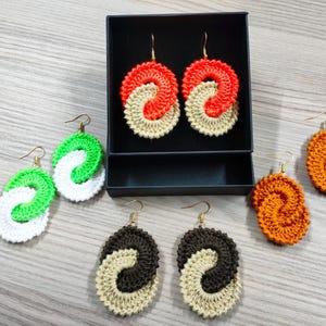 May include: A set of six crocheted earrings in various colors, including green, white, orange, brown, and beige. The earrings are in the shape of a double loop, with each loop a different color. The earrings are displayed on a black background.
