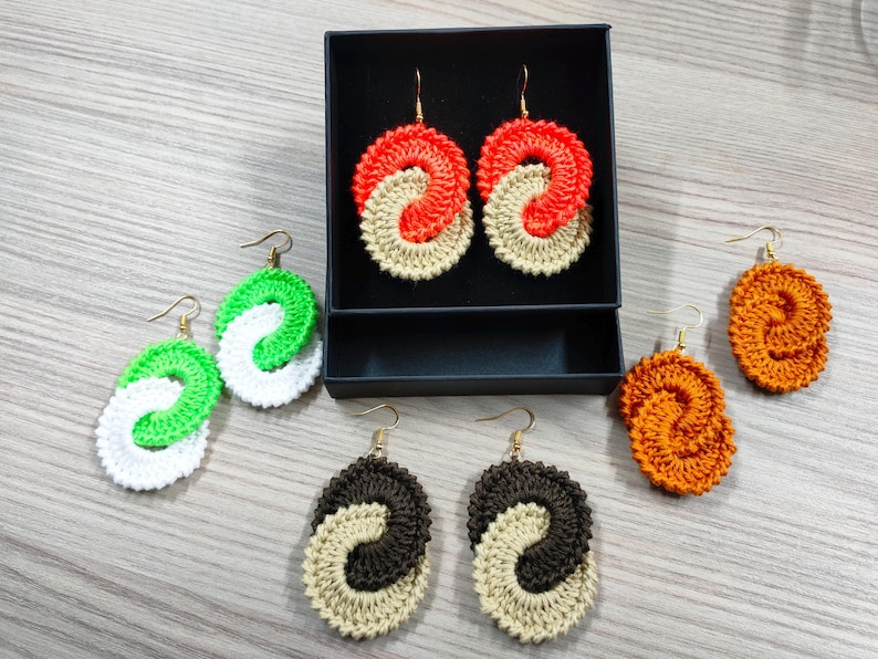 May include: A set of six crocheted earrings in various colors, including green, white, orange, brown, and beige. The earrings are in the shape of a double loop, with each loop a different color. The earrings are displayed on a black background.