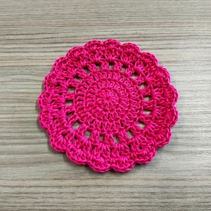 May include: A bright pink crocheted coaster with a circular design. The coaster is made of yarn and has a delicate, lacy pattern.