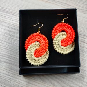 May include: A pair of crocheted earrings in a black box. The earrings are made of two colors of yarn, orange and beige, and are shaped like a figure eight.