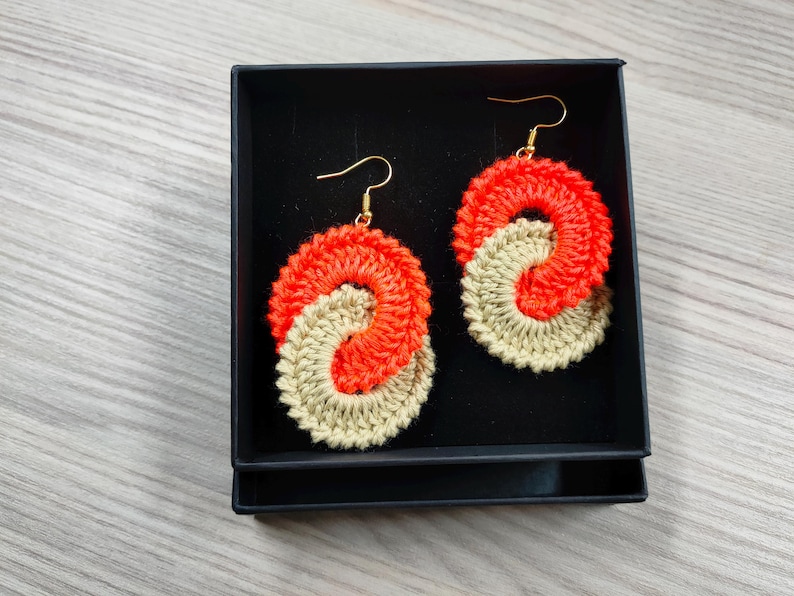 May include: A pair of crocheted earrings in a black box. The earrings are made of two colors of yarn, orange and beige, and are shaped like a figure eight.