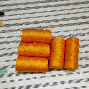 May include: Five cylindrical spools of vibrant golden-orange nylon cord arranged in a cluster. Three spools are stacked horizontally on the left, while two spools are positioned vertically side-by-side on the right, showing a braided texture.