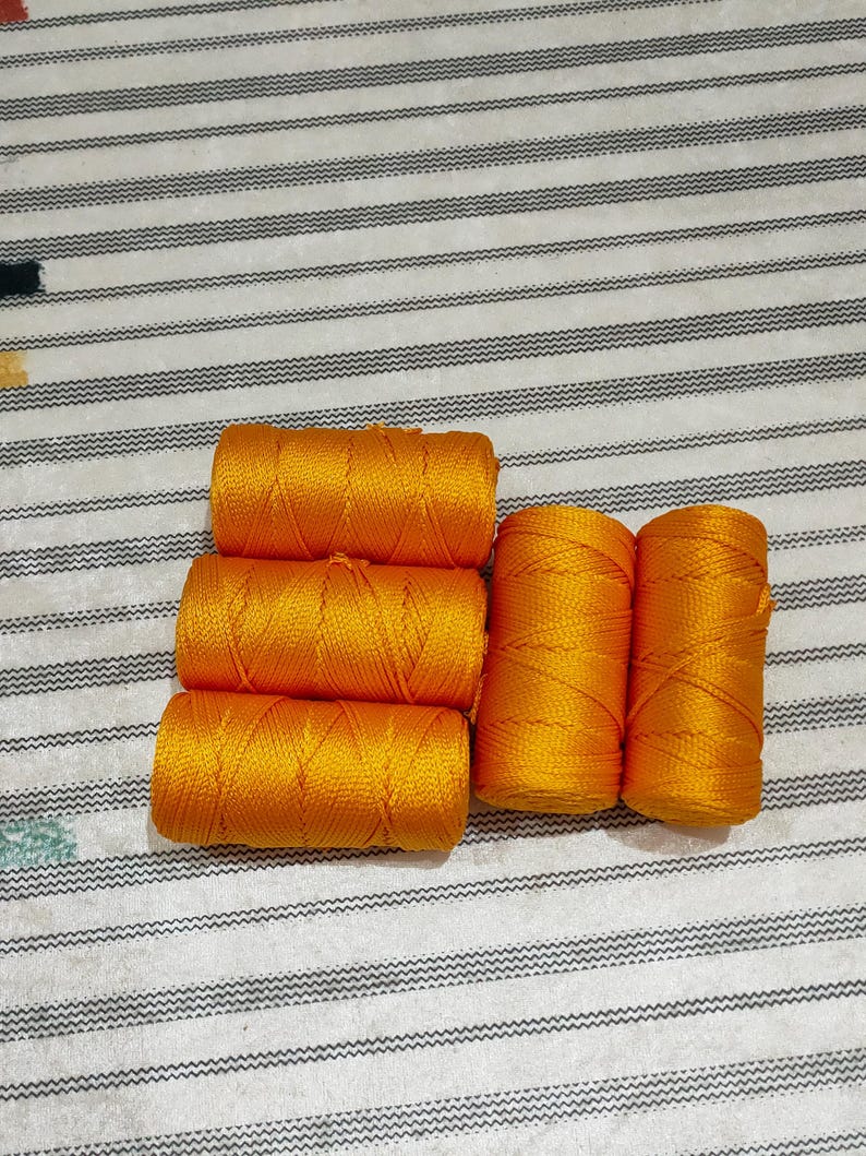 May include: Five cylindrical spools of vibrant golden-orange nylon cord arranged in a cluster. Three spools are stacked horizontally on the left, while two spools are positioned vertically side-by-side on the right, showing a braided texture.
