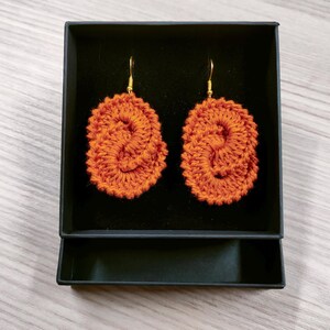 May include: A pair of orange crocheted earrings in a black box. The earrings are oval shaped and have a twisted design.