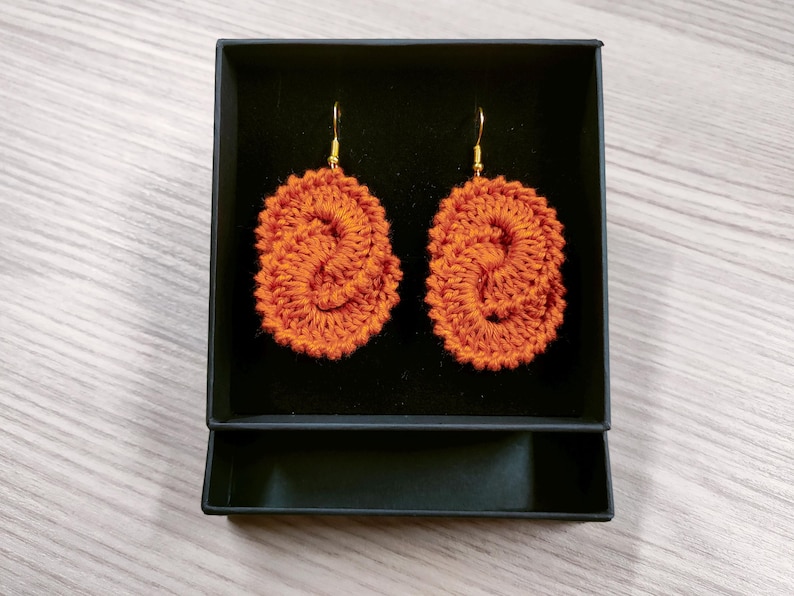 May include: A pair of orange crocheted earrings in a black box. The earrings are oval shaped and have a twisted design.