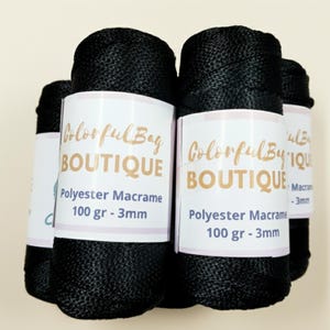 May include: Several spools of black polyester macrame cord, each wrapped in a white paper label featuring the text Colorful Bag BOUTIQUE, Polyester Macrame, 100 gr - 3mm. The cord has a smooth, braided texture and a consistent dark black color.