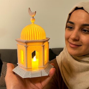 Puede incluir: Una linterna decorativa amarilla y gris con una parte superior en forma de cúpula y un remate de media luna. La linterna tiene ventanas arqueadas y una vela encendida en el interior. La linterna está en la mano de una persona.