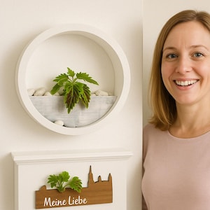 May include: A white wall with a round wooden frame with a green plant inside. Below the frame is a wooden sign with the text "Meine Liebe" and a small green plant.