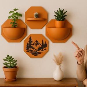 Peut inclure: Trois pots muraux hexagonaux orange avec un motif de scène de montagne noire. Les pots sont fixés à un mur blanc. Une femme pointe du doigt le pot avec la scène de montagne.