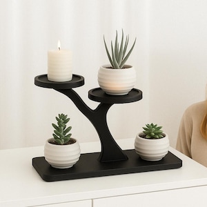 Op de afbeelding: Een zwarte, drietraps plantenstandaard. De bovenste trede heeft een witte kaars, de middelste trede heeft een kleine succulent in een pot en de onderste trede heeft een grotere succulent in een pot.