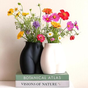 May include: Two body-shaped vases, one black and one white, hold colorful flowers. The vases sit atop two books with the titles "Botanical Atlas" and "Visions of Nature". The flowers include yellow, pink, and red blooms.