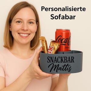 Könnte beinhalten: Ein grauer Snackhalter mit dem Text "Snack Bar Mattis" an der Seite. Der Halter hat Fächer für eine Dose und zwei Snackriegel. Eine Dose Coca-Cola Zero befindet sich im Halter.