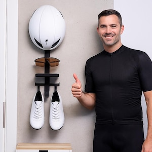 Könnte beinhalten: Ein weißer Fahrradhelm, eine Sonnenbrille und weiße Radschuhe werden auf einem schwarzen Wandregal präsentiert. Das Regal ist für die Aufbewahrung von Fahrradausrüstung konzipiert. Eine Person in schwarzer Sportkleidung gibt ein Daumen hoch.