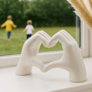 May include: A white ceramic sculpture of two hands forming a heart shape. The sculpture sits on a white windowsill, with a blurred background of a green lawn and two children playing. A vase of dried pampas grass is to the right.