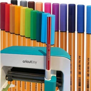 Op de afbeelding: Een Cricut Joy snijmachine met een rode Stabilo Pen 68 erin gestoken. De machine is wit met een turquoise bovenkant en een zwarte snijmes. De machine is omringd door een verscheidenheid aan gekleurde Stabilo pennen.