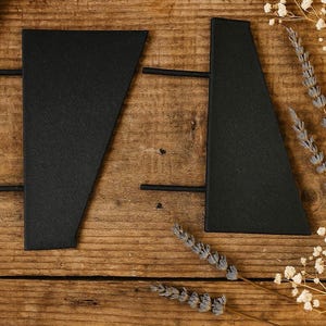 Puede incluir: Dos objetos negros de forma geométrica con barras horizontales, posiblemente para un proyecto de manualidades, están dispuestos sobre una superficie de madera rústica. Trigo seco y delicadas flores blancas añaden un toque natural.