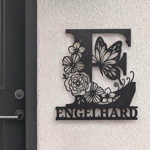 Peut inclure: Panneau de monogramme en métal noir avec la lettre "E", un papillon et des motifs floraux. Le nom "ENGELHARD" est affiché en dessous. Le panneau est fixé sur un mur texturé blanc cassé.