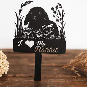 Könnte beinhalten: Schwarzer Metall-Gartenstecker mit einer Silhouette eines Kaninchens, umgeben von Blumen und Blättern. Der Stecker trägt die Aufschrift "I ❤️ My Rabbit" in Weiß und Silber. Der Stecker steht auf einer Holzoberfläche.