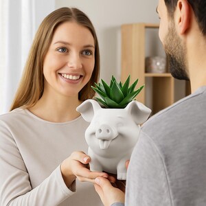 Puede incluir: Una maceta de cerámica blanca con forma de cerdo que contiene una planta suculenta verde. La maceta de cerdo tiene una cara sonriente con la lengua fuera. La maceta está siendo sostenida por una persona.