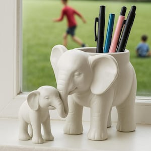 Op de afbeelding: Een witte keramische pennenhouder in de vorm van een olifant en een kleiner bijpassend olifantenbeeldje. De pennenhouder heeft een groot lichaam en grote oren en bevat meerdere pennen. De pennen zijn zwart, blauw en roze. De achtergrond is een vage buitenscène.