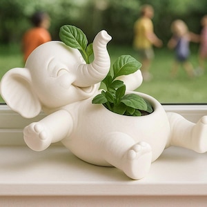 Könnte beinhalten: Ein cremefarbener Elefanten-Pflanztopf aus Keramik, der lächelt und grüne Pflanzen hält. Der Elefant liegt, mit erhobenem Rüssel und ausgestreckten Beinen. Der Pflanztopf steht auf einer weißen Oberfläche, mit anderen Pflanzen im Hintergrund.