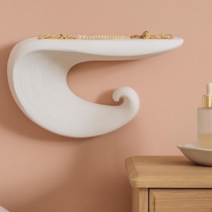 Può includere: Una mensola a muro bianca a forma di onda con superficie strutturata, con gioielli dorati. Due bottiglie di vetro con tappi dorati e bianchi sono su un piccolo piatto bianco su un comodino di legno. Il muro è color pesca.