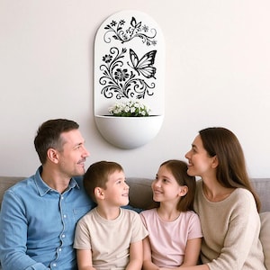 Peut inclure: Un pot mural blanc avec un motif de papillon noir et de fleurs. Le pot est de forme ovale et a une lèvre blanche. Une famille de quatre personnes est assise sur un canapé devant le pot.