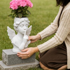 May include: A white angel-shaped planter with a finger to its lips, holding pink roses. The angel planter has detailed wings and is placed on a gray stone base. The flowers add a pop of color to the serene scene.