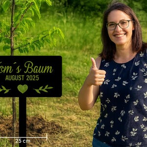 Könnte beinhalten: Ein schwarzes Schild mit dem Text "Tom's Baum AUGUST 2025" in grünen Buchstaben, mit einem grünen Herz und Blättermuster, steht vor einem jungen Baum. Das Schild ist 25 cm breit. Eine Person gibt ein Daumen hoch.