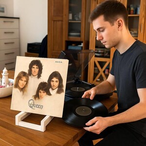 May include: A white wooden stand displaying a Queen album cover featuring band portraits. A black vinyl record sits on a turntable, with another held close. The album cover has the word "Queen" in a stylized font.