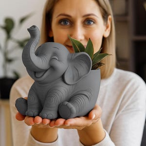 Puede incluir: Un macetero gris con forma de elefante sonriente, que sostiene una pequeña planta verde. El elefante tiene grandes orejas y una trompa enroscada. El macetero es de material liso y es un objeto decorativo encantador.