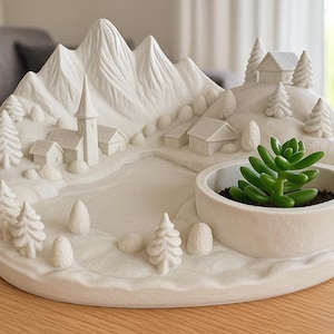 Può includere: Una fioriera in ceramica bianca a forma di villaggio di montagna, con una chiesa, case, alberi e un lago. Una piccola pianta succulenta è in un vaso rotondo a destra. La fioriera è su un tavolo di legno marrone chiaro.