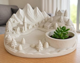 Op de afbeelding: Een witte keramische plantenbak in de vorm van een bergdorp, met een kerk, huizen, bomen en een meer. Een kleine vetplant staat in een ronde pot aan de rechterkant. De plantenbak staat op een lichtbruine houten tafel.