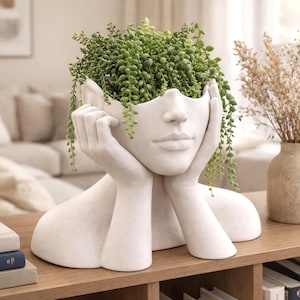 Puede incluir: Una maceta de cerámica blanca con forma de rostro, con las manos sosteniendo la cabeza, llena de plantas verdes colgantes. La pieza decorativa está sobre una estantería de madera, con otra decoración.