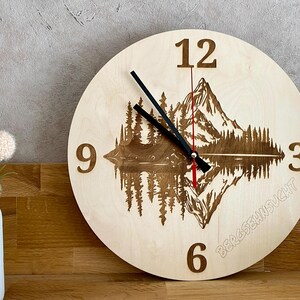 Op de afbeelding: Een houten klok met een gegraveerd berg- en meerlandschap op de wijzerplaat. De klok heeft zwarte wijzers en Romeinse cijfers. De klok staat op een houten oppervlak.