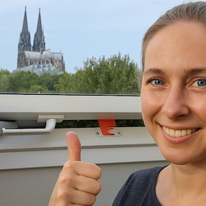 Può includere: Un telaio di finestra bianco con una maniglia bianca e un fermo di sicurezza arancione. Lo sfondo presenta una grande cattedrale con due guglie e alberi verdi sotto un cielo nuvoloso.