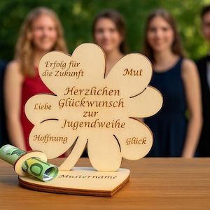 Op de afbeelding: Een houten klavertjevier met de woorden "Erfolg für die Zukunft", "Liebe", "Hoffnung", "Mut", "Glück", "Herzlichen Glückwunsch zur Jugendweihe" en "Mustername" erop gegraveerd. Het klavertjevier staat op een houten tafel met een opgerold biljet in de basis.