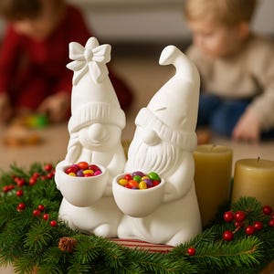 以下が含まれることがあります： カラフルなキャンディーが入った小さなボウルを持った、白いセラミック製のノームの置物2つ。ノームは休日の装飾の一部で、赤いベリーとキャンドルを背景に、緑のベッドの上に座っています。