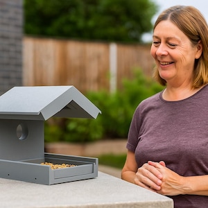 May include: A grey wooden bird feeder with a pitched roof and a circular aperture. The feeder is filled with birdseed and sits on a concrete surface. A woman in a purple top smiles in the background.