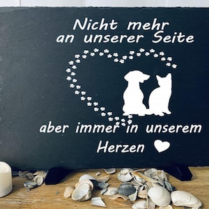 Op de afbeelding: Zwarte leisteenplaat met witte potafdrukken die een hartvorm vormen. Binnen het hart bevinden zich silhouetten van een hond en een kat. De tekst luidt "Niet meer aan onze zijde, maar altijd in ons hart" met een wit hart symbool.